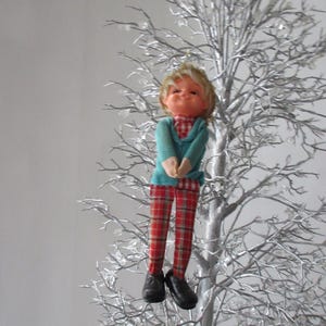 May include: A doll with blonde hair and a smiling face, wearing a teal long-sleeved top over a red and white checkered shirt, paired with red plaid trousers and black shoes, suspended on a silver-colored decorative tree with clear accents.