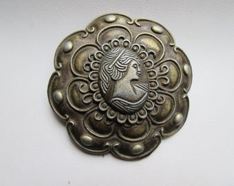 Könnte beinhalten: Antik-Stil Brosche mit einem Cameo-Design. Die Brosche zeigt ein zentrales ovales Cameo-Profil einer Frau, umgeben von einem verzierten, blütenförmigen Rahmen mit Bronze-Finish. Ein vintage-inspiriertes Accessoire.