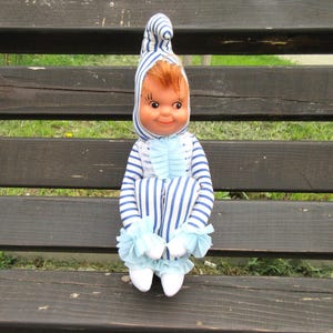 Könnte beinhalten: Eine Vintage-Puppe mit lächelndem Gesicht, die einen blau-weiß gestreiften Pyjama und eine passende Mütze trägt. Die Puppe hat rotes Haar und weiße Schuhe. Der Pyjama hat hellblaue Rüschen an den Handgelenken und Knöcheln. Die Puppe sitzt auf einer Holzbank.