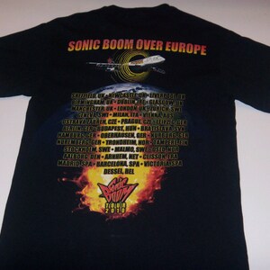 K&ouml;nnte beinhalten: Schwarzes T-Shirt mit einem Grafikdruck eines Flugzeugs, das &uuml;ber einem Globus fliegt, und dem Text "Sonic Boom Over Europe". Das Shirt listet auch die europ&auml;ischen St&auml;dte auf, in denen die Band auf ihrer Tour 2010 auftreten wird.