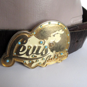 Może przedstawiać: Złota metalowa klamra do paska z napisem Elvis Las Vegas czcionką odręczną na kremowym tle, przedstawiająca sylwetkę artysty. Klamra jest ozdobiona małymi turkusowymi ćwiekami i przymocowana do ciemnobrązowego skórzanego paska.
