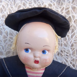 May include: A doll with a smooth, pale face, bright blue eyes, and painted red lips. The doll wears a black fabric beret-style hat and a black collared garment over a pink and white horizontally striped shirt, with light blonde hair visible.