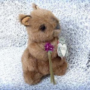 May include: A brown plush squirrel toy with soft fur, dark eyes, and pointed ears. The squirrel holds a small purple flower with a green ribbon stem. A heart-shaped tag featuring a grey bear illustration and the text GUND is attached to the side.