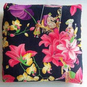 May include: A black pillow with a floral pattern featuring pink lilies, yellow orchids, and purple flowers. The pillow has a dark background with a repeating pattern of people in ornate clothing.