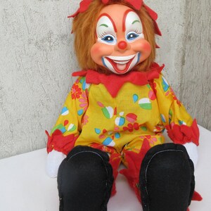 May include: A clown doll with orange hair, a painted face, and a wide smile. The doll wears a yellow jumpsuit featuring a colourful floral pattern, red ruffled collar and cuffs, white fabric hands, and large black felt shoes with red trim.