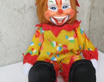 May include: A clown doll with orange hair, a painted face, and a wide smile. The doll wears a yellow jumpsuit featuring a colorful floral pattern, red ruffled collar and cuffs, white fabric hands, and large black felt shoes with red trim.