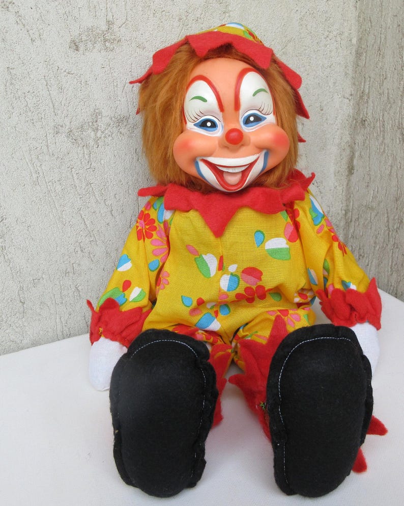 May include: A clown doll with orange hair, a painted face, and a wide smile. The doll wears a yellow jumpsuit featuring a colourful floral pattern, red ruffled collar and cuffs, white fabric hands, and large black felt shoes with red trim.