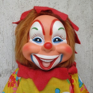May include: A clown doll with a moulded plastic face, painted features including a red nose, wide smile, and blue accents. The doll has synthetic brown hair, a red fabric collar, and a yellow shirt with a colourful floral pattern.