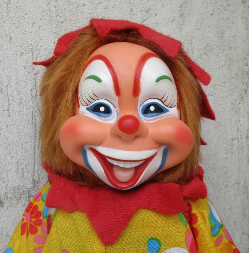May include: A clown doll with a moulded plastic face, painted features including a red nose, wide smile, and blue accents. The doll has synthetic brown hair, a red fabric collar, and a yellow shirt with a colourful floral pattern.