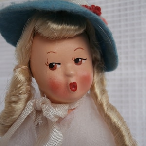 May include: A porcelain doll with blonde hair styled in pigtails, wearing a blue hat with red flowers and a white dress.