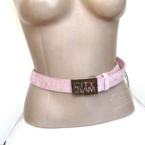 Könnte beinhalten: Ein hellrosa Gürtel mit einer silbernen Schnalle. Auf der Schnalle steht "CITY GLAM". Der Gürtel wird auf einer Schaufensterpuppe präsentiert.