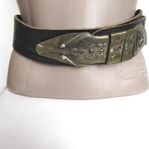 Peut inclure: Une ceinture en cuir noir dotée d'une large sangle texturée et d'une grande boucle en métal doré vieilli. La boucle a la forme d'une tête stylisée et est ornée de petits strass incrustés de couleurs rouge, orange, verte et transparente.