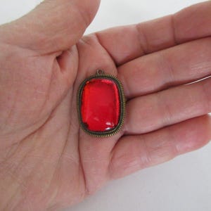 Op de afbeelding: Een rechthoekige, rode hanger met een bronzen, sierlijke lijst. De hanger wordt in de hand van een persoon gehouden. De rode steen heeft een glanzende afwerking. Het frame heeft een kleine lus aan de bovenkant voor een ketting.