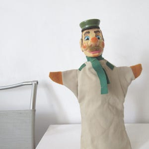 May include: A hand puppet featuring a painted wooden head with a green cap, blue eyes, and a mustache. The puppet wears a beige fabric body with orange sleeve ends and a green felt scarf tied around the neck, creating a classic character design.