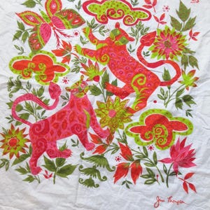 May include: A white fabric square featuring a vibrant pattern of two stylized pink and orange leopards, a large butterfly, green and red floral motifs, and abstract cloud shapes. The signature Jim Thompson is printed in red in the bottom corner.