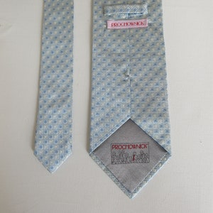 May include: A light blue necktie with a repeating square pattern in white and light blue. The back of the tie features a pink label with the text "PROCHOWNICK" and a gray lining with a small illustration and the brand name.