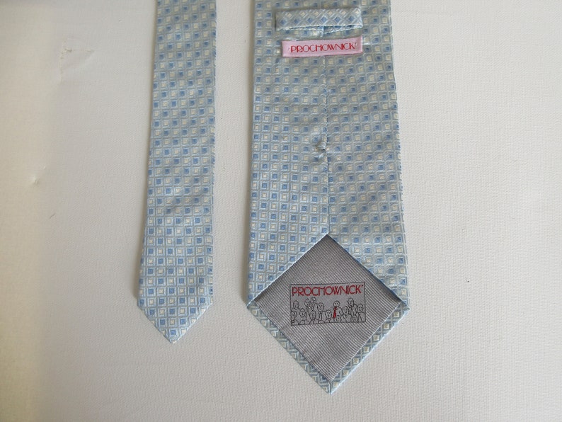 May include: A light blue necktie with a repeating square pattern in white and light blue. The back of the tie features a pink label with the text "PROCHOWNICK" and a gray lining with a small illustration and the brand name.