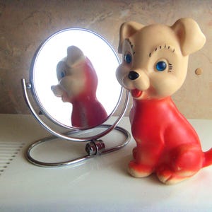 May include: A small, moulded rubber dog figurine with a cream-coloured head and a bright red body. The dog has blue eyes, a black nose, and a red tongue. Beside the figurine stands a small, round, silver-toned metal vanity mirror on a stand.