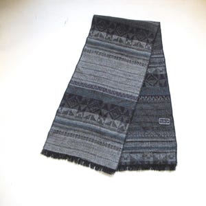 May include: A gray and blue patterned scarf with a fringed edge. The scarf features geometric designs and horizontal stripes in shades of gray, blue, and black. A small label is visible on the scarf. The scarf is made of a soft material.