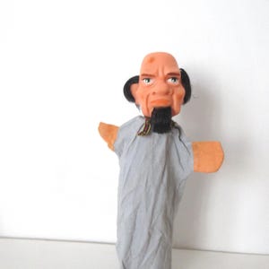 May include: A hand puppet featuring a molded rubber head with a bald scalp, a small black goatee, and black hair on the sides. The puppet wears a light gray fabric robe with tan fabric hands extending from the sleeves, creating a character figure.