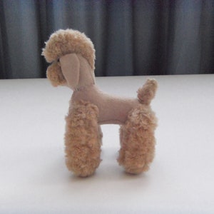 May include: A small, beige plush toy poodle with curly fur. The toy is standing on a white surface.