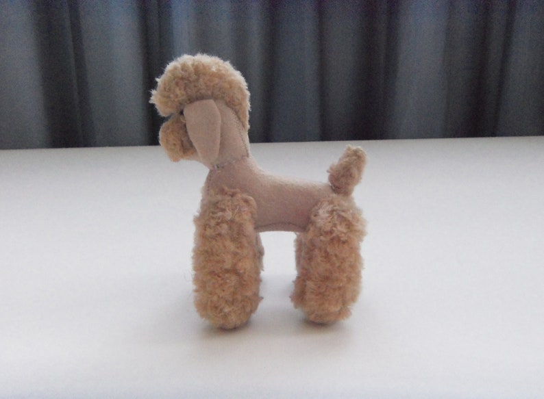 May include: A small, beige plush toy poodle with curly fur. The toy is standing on a white surface.