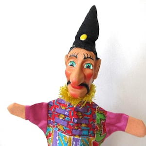 May include: A hand puppet featuring a character with a pointed black hat, a yellow button, and a yellow fringe collar. The puppet has a painted face with a long nose, black moustache, and green eyes, wearing a colourful patterned shirt with purple sleeves.