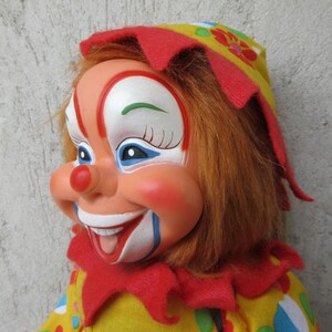 May include: A clown doll with a painted face featuring a red nose, white mouth area, and blue eye accents. The doll has orange synthetic hair, a red felt collar, and a yellow hat and outfit decorated with colourful polka dots and floral patterns.