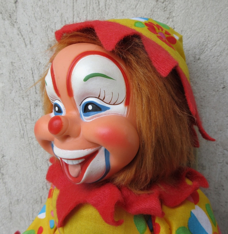 May include: A clown doll with a painted face featuring a red nose, white mouth area, and blue eye accents. The doll has orange synthetic hair, a red felt collar, and a yellow hat and outfit decorated with colourful polka dots and floral patterns.