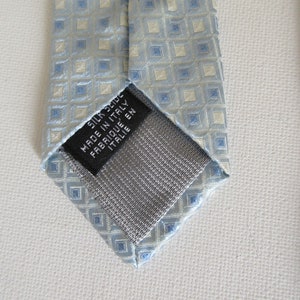 May include: A light blue silk tie with a repeating square pattern in light blue and off-white. The tie has a gray backing and a black label that reads "SILK 100% MADE IN ITALY FABRIQUE EN ITALIE".