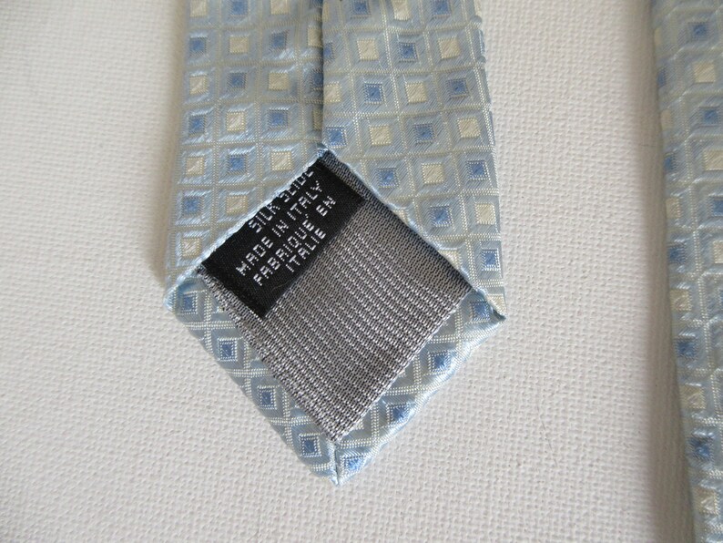 May include: A light blue silk tie with a repeating square pattern in light blue and off-white. The tie has a gray backing and a black label that reads "SILK 100% MADE IN ITALY FABRIQUE EN ITALIE".