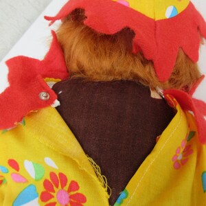 May include: A plush doll with orange hair and a yellow fabric outfit featuring a colourful floral pattern. The doll wears a red felt collar with pointed edges and a matching red felt hat. The garment is secured with a small silver metal snap.