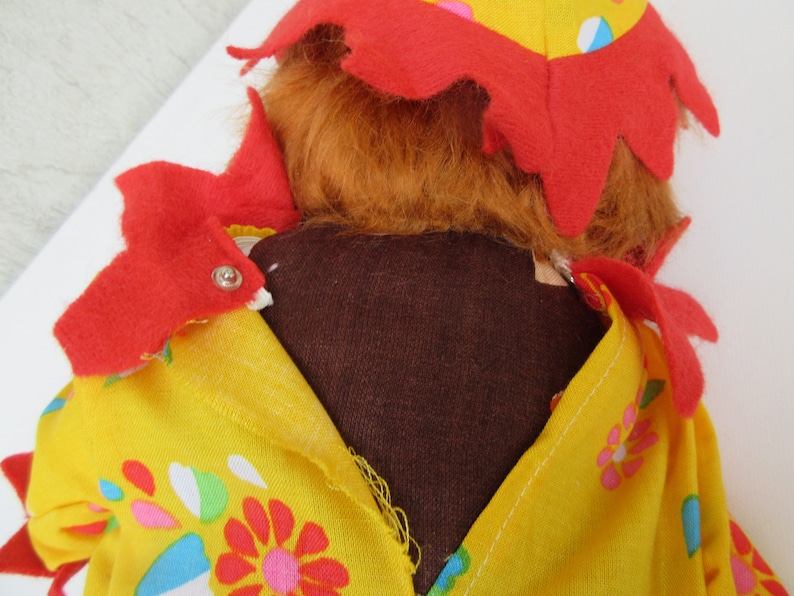 May include: A plush doll with orange hair and a yellow fabric outfit featuring a colourful floral pattern. The doll wears a red felt collar with pointed edges and a matching red felt hat. The garment is secured with a small silver metal snap.