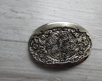 Könnte beinhalten: Eine ovale Gürtelschnalle aus silberfarbenem Metall mit einem komplexen Prägemuster. Das Design zeigt ein zentrales kreisförmiges Motiv mit wirbelnden Filigrandetails, das von floralen und blattartigen Gravuren auf der Oberfläche umgeben ist.