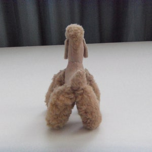 May include: A plush toy poodle in a light brown color. The toy is sitting on a white surface and is facing away from the camera.