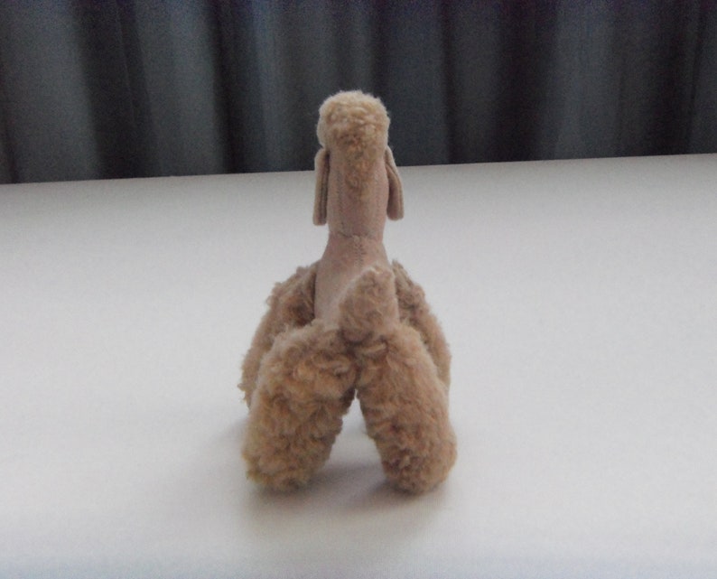 May include: A plush toy poodle in a light brown color. The toy is sitting on a white surface and is facing away from the camera.