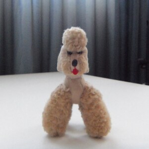 May include: A small, beige, plush toy poodle with black eyes and a red mouth. The toy is standing on a white surface.