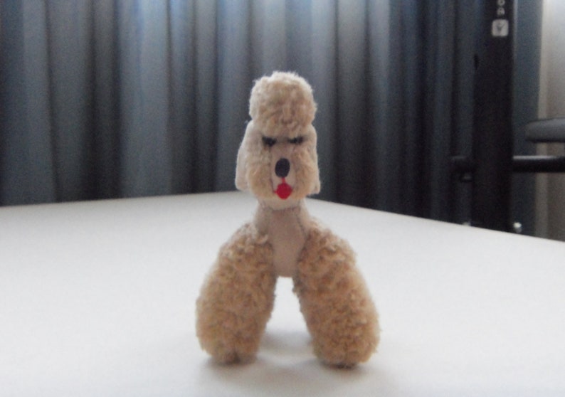 May include: A small, beige, plush toy poodle with black eyes and a red mouth. The toy is standing on a white surface.