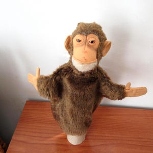 May include: A brown plush monkey hand puppet featuring a soft, fuzzy body, a light tan face with a white chin patch, and moulded plastic hands. The puppet has a neutral expression with dark eyes and large ears, designed for interactive play.