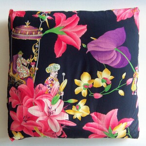 May include: A black pillow with a floral pattern. The pattern includes pink, purple, yellow, and green flowers. There are also two figures in the pattern, one wearing a turban and the other wearing a crown.