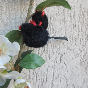 May include: A handmade black plush devil doll with red horns and accents, perched on a branch with green leaves and white flowers. The doll has a small tail and is set against a textured, light gray wall.