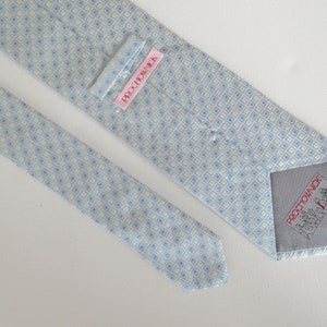 May include: A light blue and cream-colored tie with a repeating square pattern. The tie has a label that reads "PROCHOWNICK" in red text. The tie is made of a smooth fabric.