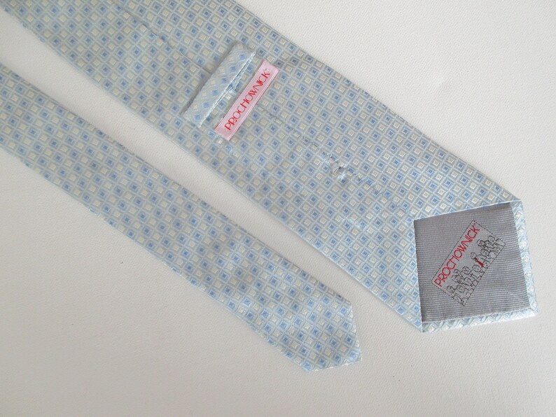 May include: A light blue and cream-colored tie with a repeating square pattern. The tie has a label that reads "PROCHOWNICK" in red text. The tie is made of a smooth fabric.