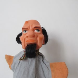 Peut inclure: Marionnette à main avec une tête chauve, une expression sévère et une barbe noire. La marionnette a des cheveux noirs sur les côtés de la tête et porte une chemise grise avec des manches orange. Le visage de la marionnette est de couleur pêche clair.