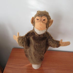 May include: A vintage monkey puppet with a brown fur body, tan face, and outstretched arms. The puppet has large ears and a white collar. It is displayed on a wooden surface.