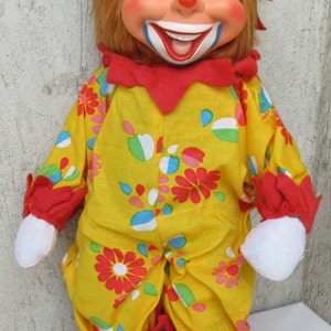 May include: A clown doll with a painted face, orange hair, and a red hat. It wears a yellow jumpsuit featuring a colourful floral and teardrop pattern, red ruffled cuffs and collar, white fabric hands, and black shoes with red ruffled trim.
