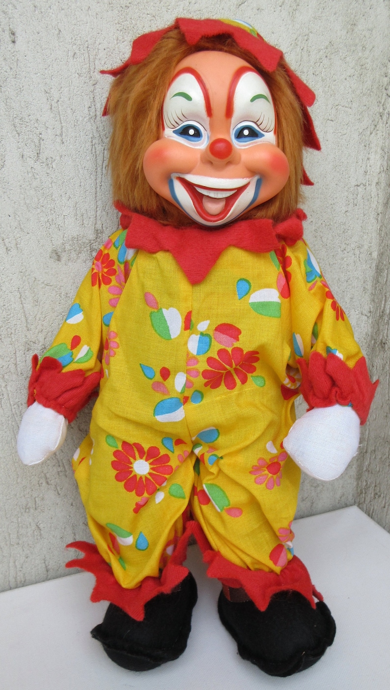 May include: A clown doll with a painted face, orange hair, and a red hat. It wears a yellow jumpsuit featuring a colourful floral and teardrop pattern, red ruffled cuffs and collar, white fabric hands, and black shoes with red ruffled trim.