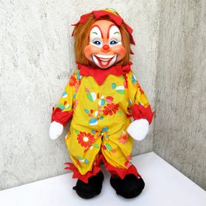 May include: A clown doll with orange hair, a painted face featuring a wide smile, and a yellow jumpsuit decorated with red, pink, and blue floral patterns. The outfit includes red ruffled collars and cuffs, white gloves, and black shoes.