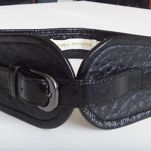 May include: A black leather belt with a buckle and a textured surface. The belt has a label that reads "ruuhe simonsen".