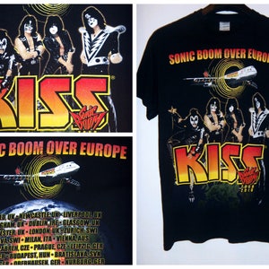 K&ouml;nnte beinhalten: Schwarzes T-Shirt mit einem Grafikdruck der Band KISS und einem Flugzeug mit dem Text "Sonic Boom Over Europe". Die Bandmitglieder tragen ihr charakteristisches Make-up und ihre Kost&uuml;me. Der Text "KISS" ist in gro&szlig;en roten, gelben und orangen Buchstaben. Der Text "Sonic Boom Over Europe" ist in wei&szlig;en Buchstaben.